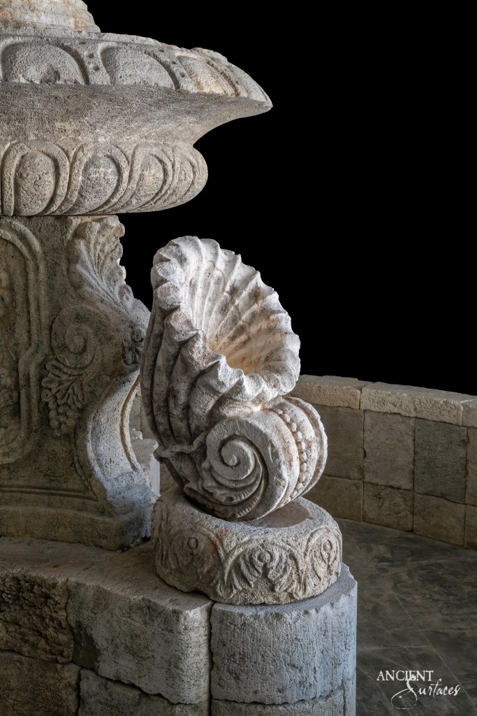 Ancient Greek-style limestone fountain by Ancient Surfaces, adorned with intricate shell motifs and other carvings, reflecting the rich artistic heritage of classical Greece.