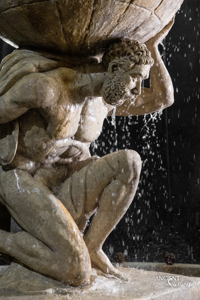 Close-up view of a vintage limestone fountain by Ancient Surfaces, featuring a detailed sculpture of a man holding a sphere over his head, with water cascading down the structure.